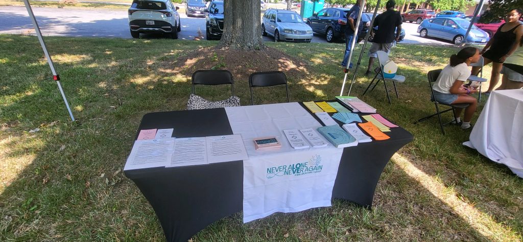 Outreach table with black tableclotth, Never Alone Never Again banner, and covered with various posters and pamphlets