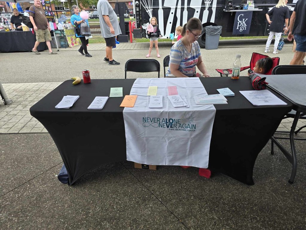 Photo of Nar-Anon outreach table with black tablecloth, a banner showing the "Never Alone Never Again"  imagery, and outreach pamphlets.